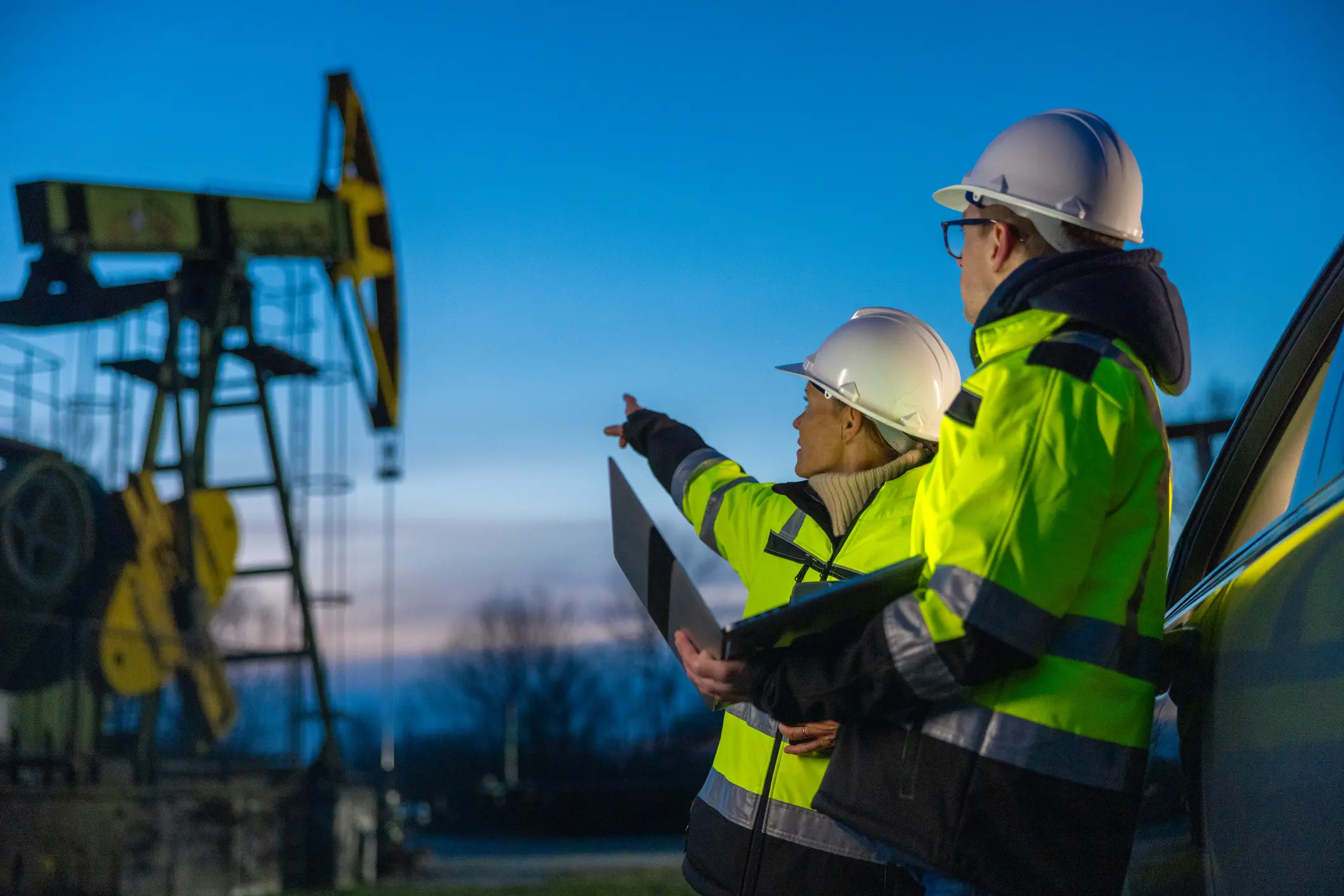 Female engineer pointing at machinery while planning with partner over laptop by car at field during sunset
